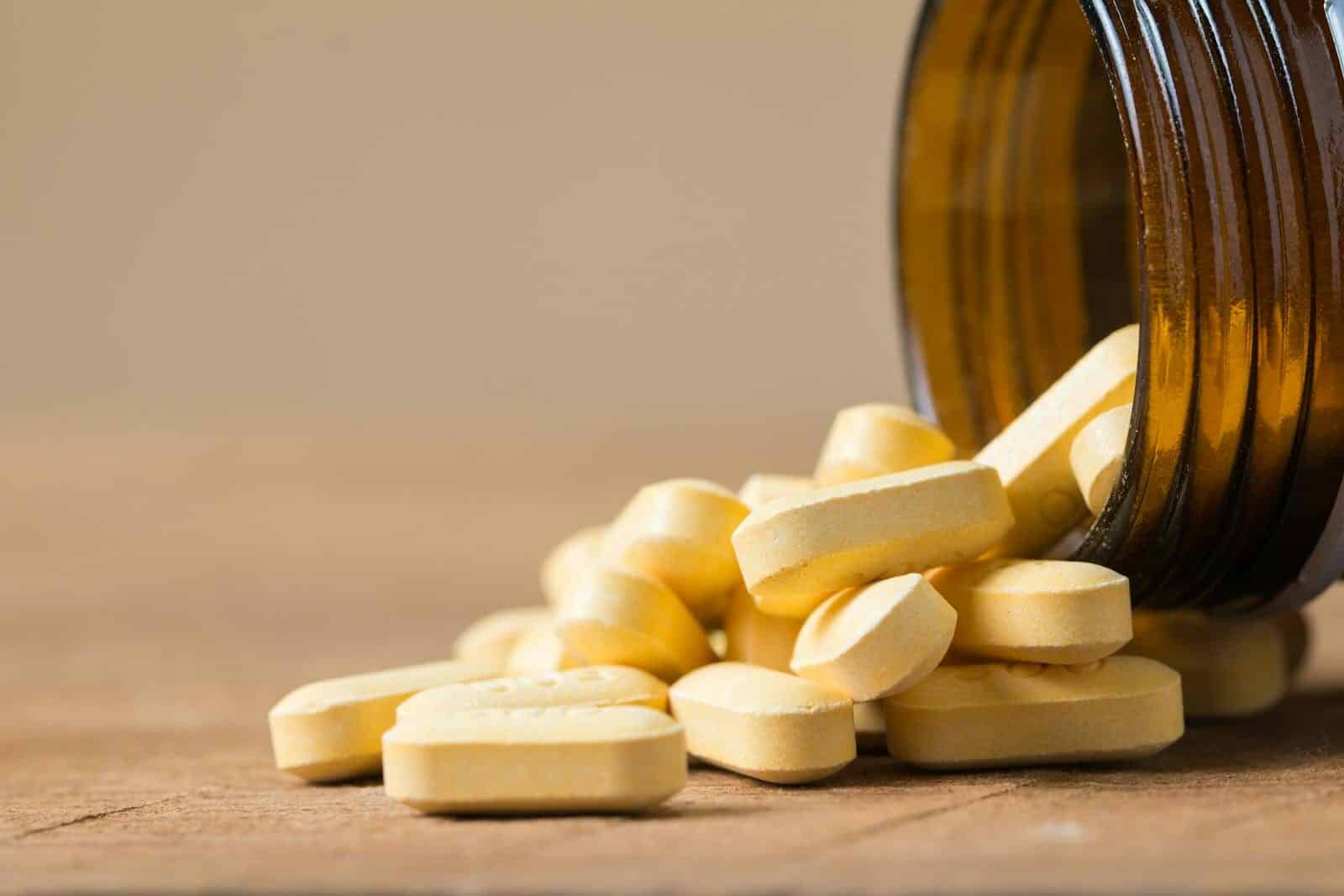 A close-up image of vitamin tablets spilling out of a transparent brown bottle on a wooden surface.